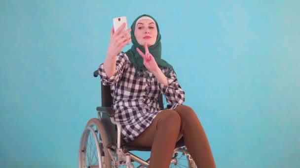 Mladá muslimka žena se zdravotním postižením na invalidním vozíku se selfie se svým telefonem na modrém pozadí — Stock video