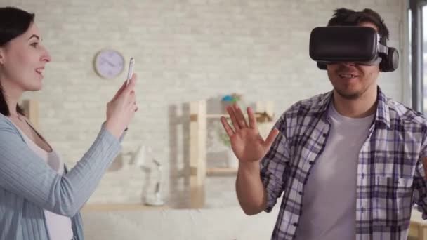 Молодая женщина снимает реакцию мужчины в очках виртуальной реальности по телефону — стоковое видео
