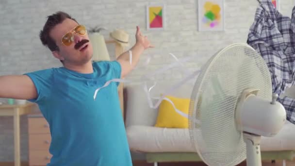 Grappige jonge man met elektrische ventilator genieten van koele wind in zijn appartement slow mo — Stockvideo