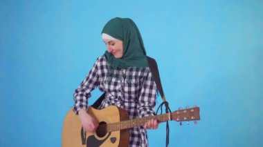 Neşeli genç Müslüman kadın açıkça mavi bir arka plan üzerinde gitar çalıyor