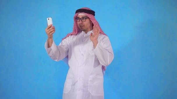 Homem árabe falando em vídeo chamada em um smartphone em um fundo azul — Vídeo de Stock
