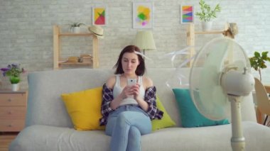 Genç bir kadın elektrikli vantilatörün önündeki koltukta oturan akıllı telefonu kullanıyor, sıcaktan kaçıyor.