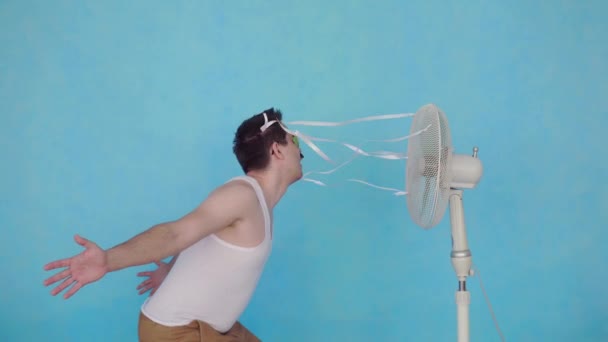 Divertido joven con ventilador eléctrico disfrutando de viento fresco sobre fondo azul — Vídeo de stock