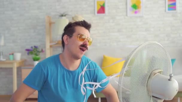 Divertido joven delante de un ventilador eléctrico de trabajo escapa del calor en el apartamento lento mo — Vídeos de Stock