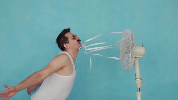 Funny young man with electric fan enjoying cool wind on blue background slow mo — Stock Video