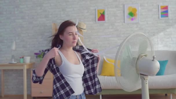 Молодая женщина, сидящая в гостиной с электрическим вентилятором — стоковое видео