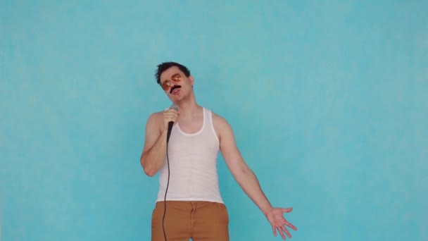 Смішний молодий чоловік в окулярах з мікрофоном в руці на синьому фоні — стокове відео