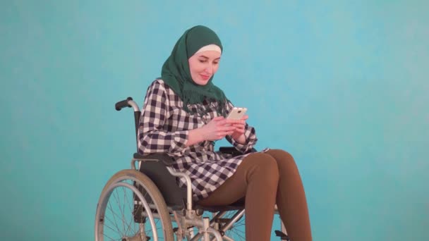 Fiatal muszlim nő fogyatékkal élő személy kerekesszékben használja a telefont a kék háttér — Stock videók