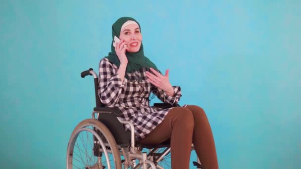 Young Muslim woman disabled in a wheelchair talking on the phone on a blue background — Stock Video