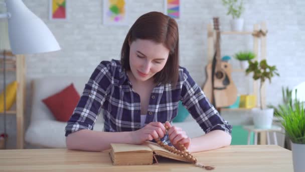 Retrato mujer joven sentada en una mesa con un rosario en las manos y leyendo un libro — Vídeos de Stock