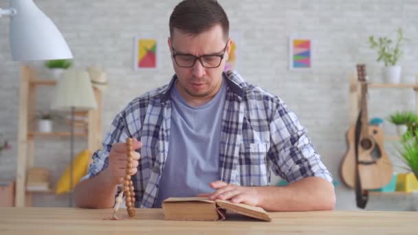Porträtt ung man med glasögon med ett Rosenkransen i händerna och läsa Bibeln — Stockvideo