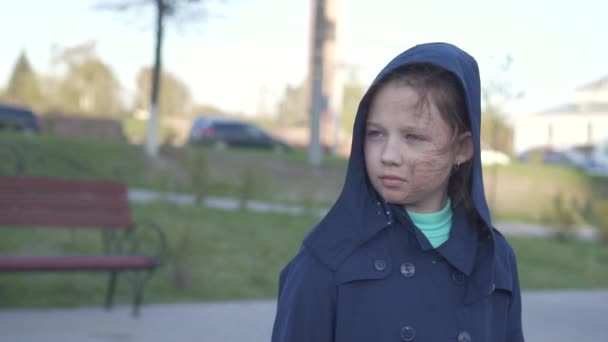 Porträtt av en sorglig tonårs flicka med en bränd ansikte gå ner på gatan med en huva på bakgrunden av stadsbilden — Stockvideo