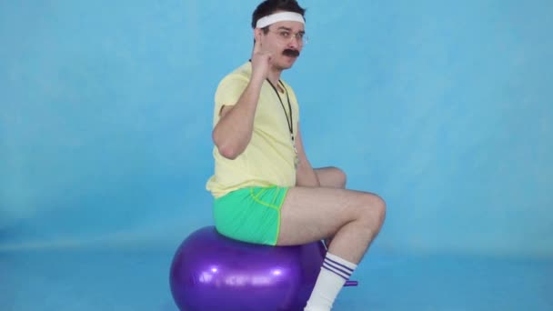 Treinador homem engraçado com um bigode dos anos 80, na bola de fitness em fundo azul slow mo — Vídeo de Stock