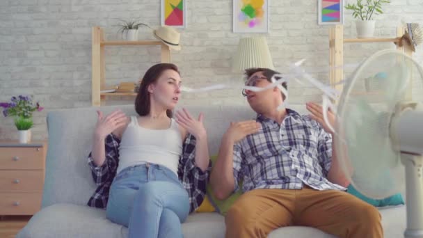 Homem e uma mulher sentados em um sofá com um ventilador elétrico estão escapando do calor lento mo — Vídeo de Stock