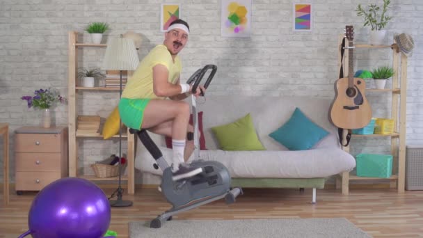 Rolig energisk man från 80-talet med en mustasch engagerad hemma på en motionscykel Slow Mo — Stockvideo