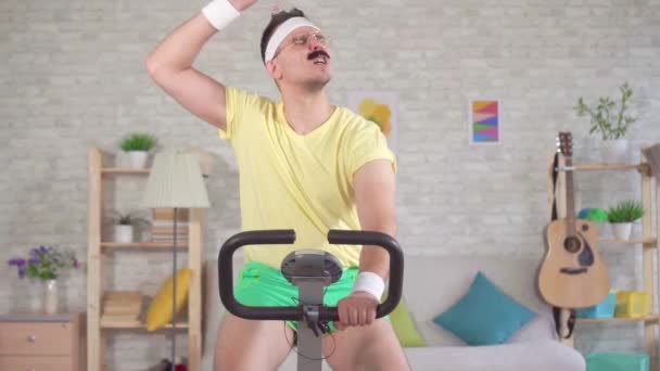 Homme drôle des années 80 avec une moustache engagé sur un vélo d'appartement et se déverse avec de l'eau de la bouteille lente mo — Video