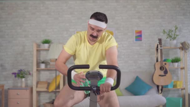 Funny sportsman from the 80s with a mustache is engaged on a exercise bike at home and drinks water — Stock Video