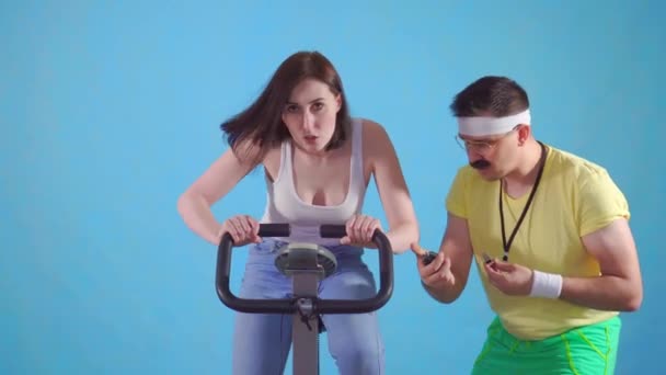 Смешной тренер 80-х с усами тренирует молодую женщину на велотренажере на синем фоне — стоковое видео
