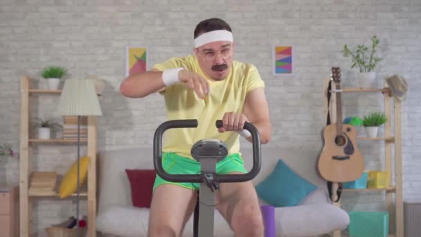 Retrato engraçado atleta homem enérgico dos anos 80 com um bigode engajado em casa em uma bicicleta de exercício slow mo — Vídeo de Stock