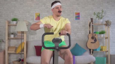 Sabit bir bisiklet üzerinde meşgul bir bıyık ile 80'li portre komik enerjik adam atlet ve yavaş mo kadar parmak gösterir