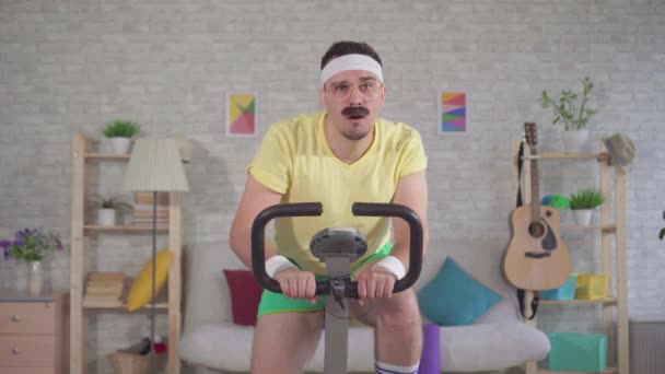 Retrato divertido atleta hombre enérgico de los años 80 con un bigote comprometido en casa en una bicicleta estacionaria — Vídeos de Stock