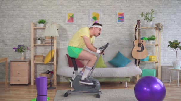 Funny man athlete from the 80s with a mustache falls off exercise bike on the couch — Stock Video