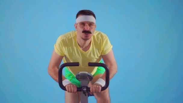 Joven atleta divertido de los años 80 con bigote en bicicleta estática sobre fondo azul — Vídeo de stock