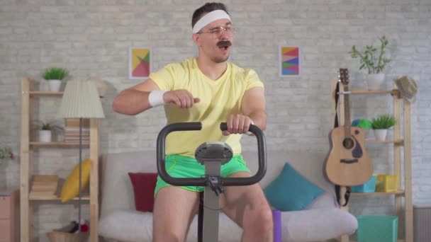 Portrait funny energetic man athlete from the 80s with a mustache engaged on a stationary bike and shows the finger up slow mo — Stock Video