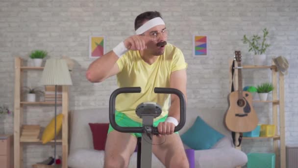 Atleta cansado divertido de los años 80 con un bigote comprometido en casa en una bicicleta de ejercicio lento mo — Vídeo de stock