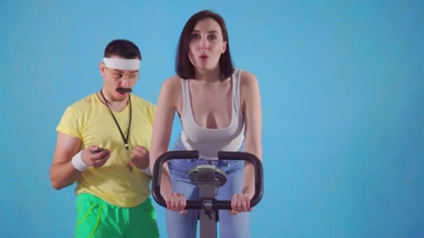 Homme drôle entraîneur des années 80 avec moustache et lunettes examine une jeune femme sur vélo d'appartement — Video