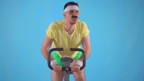 Смешной целеустремленный спортсмен 80-х с усами на велотренажере на синем фоне — стоковое видео