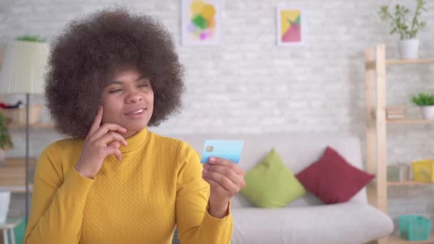 Pozitív és lelkes gyönyörű afro-amerikai nő egy afro frizura nézi a bank kártyát a kezükben a modern lakás — Stock videók