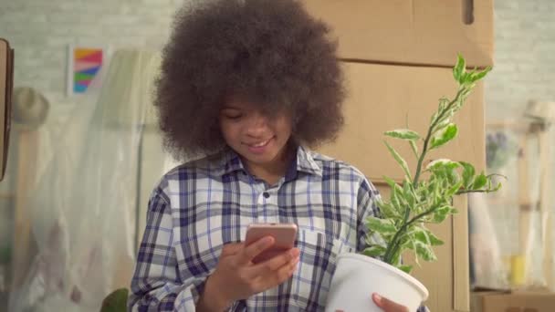 Портрет африканської жінки з рослиною в руці використовує телефон, що сидить на підлозі поруч з коробками для переміщення — стокове відео