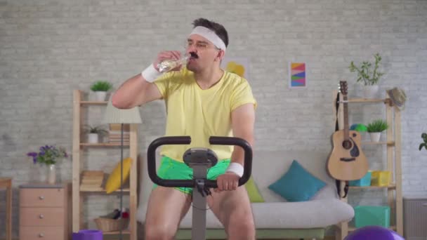 Grappige energieke atleet van de jaren 80 met een snor is bezig met een hometrainer en gietwater uit een fles op zijn hoofd — Stockvideo