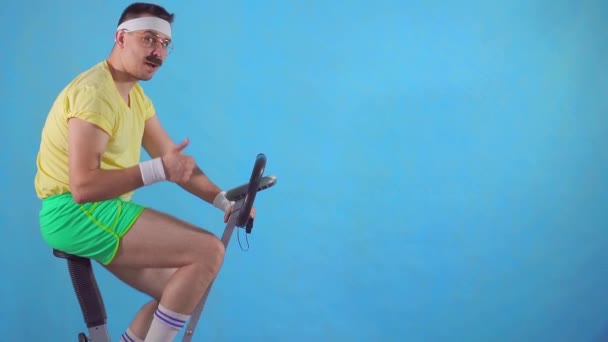 Engraçado homem dos anos 80 com um bigode em bicicleta de exercício em um fundo azul, mostra polegar-se lento mo — Vídeo de Stock