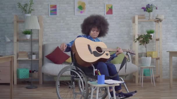 Американская женщина-инвалид с африканской прической в инвалидном кресле играет на акустической гитаре — стоковое видео