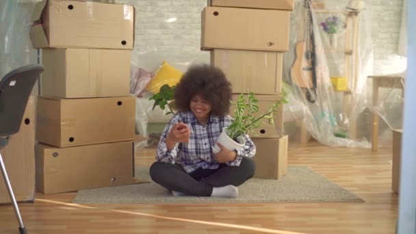 Африканська жінка з афро зачіскою з рослиною в руці використовує телефон, що сидить на підлозі поруч з коробками для переміщення — стокове відео