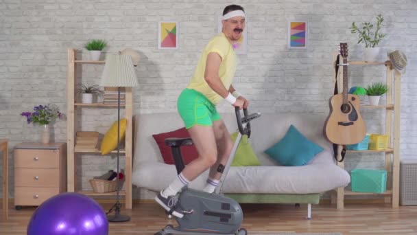 Cheerful active man athlete from the 80s with a mustache engaged at home on a exercise bike slow mo — Stock Video