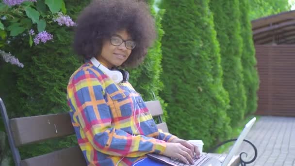 Mujer afroamericana con un peinado afro utiliza un ordenador portátil sentado en el banco en la calle mirando a la cámara — Vídeo de stock