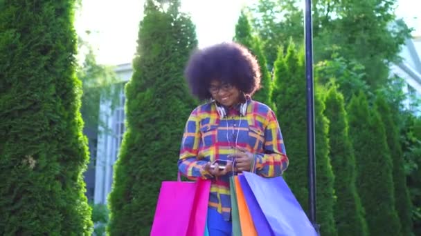 Cheerful african american woman with an afro hairstyle with packages after shopping uses a smartphone — Stock Video