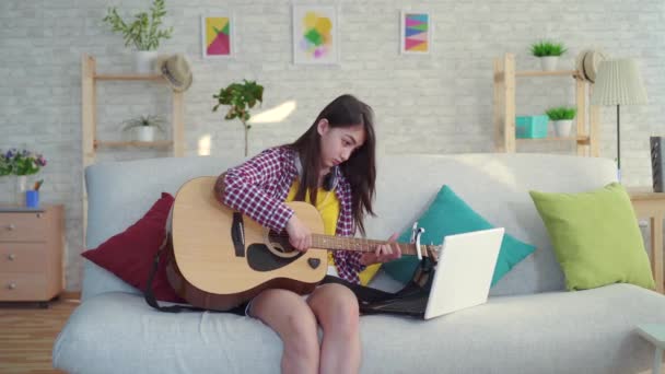 Bela menina asiática com cabelos longos na sala de estar de uma casa moderna estudando guitarra assistindo vídeos educativos na Internet em um laptop — Vídeo de Stock