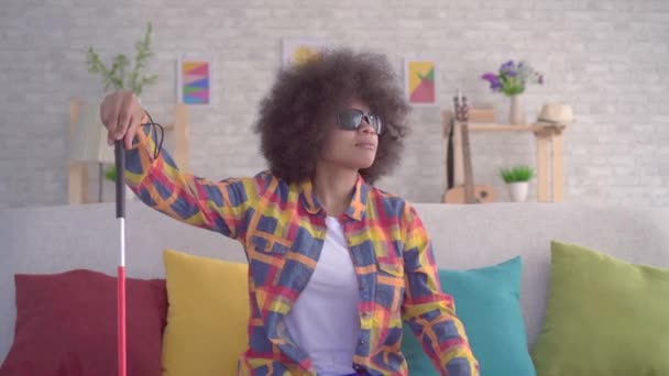 Mujer africana con un peinado afro visualmente discapacitado en gafas con un bastón en las manos sentado en el sofá — Vídeos de Stock