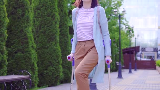 Mooie stijlvolle positieve jonge vrouw met letsel op krukken lopen door de straat zonnig — Stockvideo