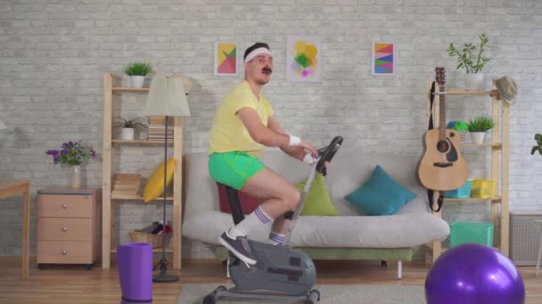 Ung rolig idrottsman från 80-talet med mustasch är engagerad på en motionscykel visar tummen upp — Stockvideo