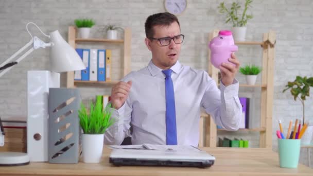 Człowiek w koszulce siedzącej na laptopie w biurze z Piggy Bank powolny mo — Wideo stockowe