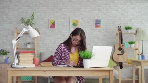 Hermosa chica asiática en gafas con el pelo largo en la sala de estar en casa con ordenador portátil y libros de texto estudiando o buscando en Internet — Vídeo de stock