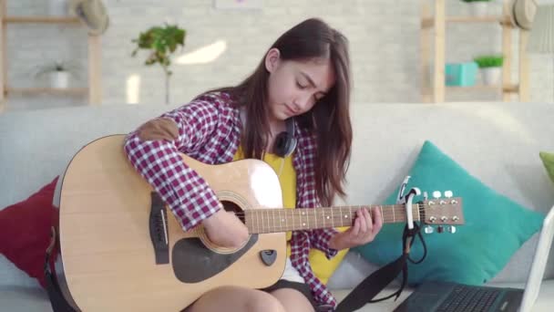 Triste belle fille asiatique avec les cheveux longs dans le salon d'une maison moderne ayant du mal à apprendre à jouer de la guitare acoustique grâce à des vidéos éducatives sur Internet — Video