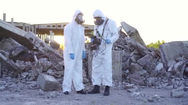 Två forskare i skyddande dräkter och masker och göra mätningar av strålning mot bakgrund av ruinerna vid solnedgången — Stockvideo