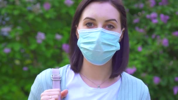 Porträtt av en ung kvinna i en medicinsk mask i bakgrunden av blommande växter i parken tittar på kameran — Stockvideo