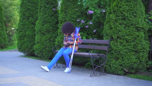 African american woman with an afro hairstyle with a broken leg with crutches sitting on a bench — Stock Video